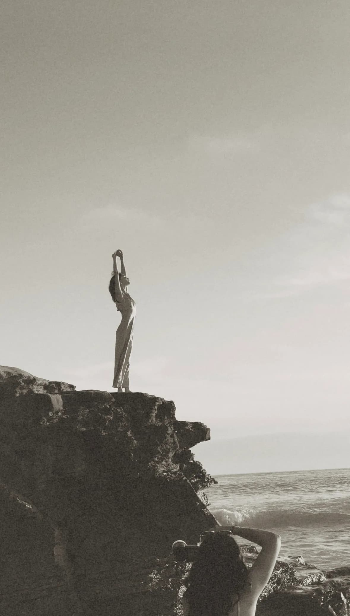 A person standing at the edge of a rocky cliff above the sea, arms stretched overhead, while someone below photographs them