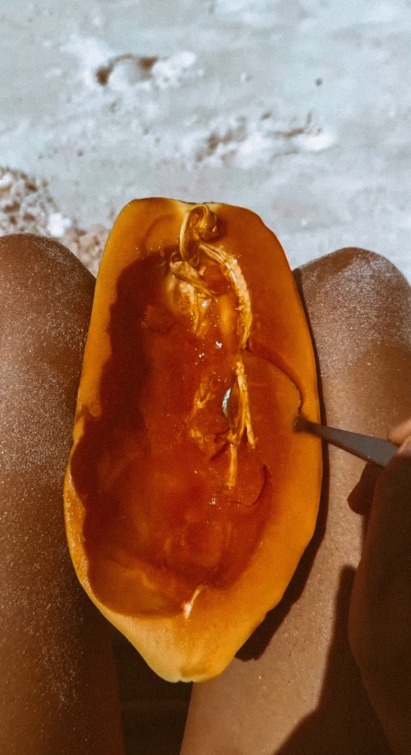 A halved papaya resting on a person's lap on the sand at the beach