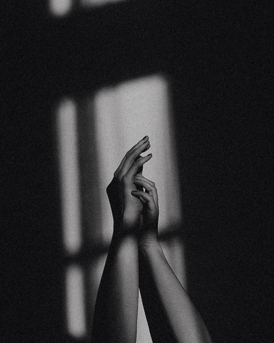 A black and white photograph of two raised hands, fingertips lightly touching, silhouetted against light from a window on a dark wall
